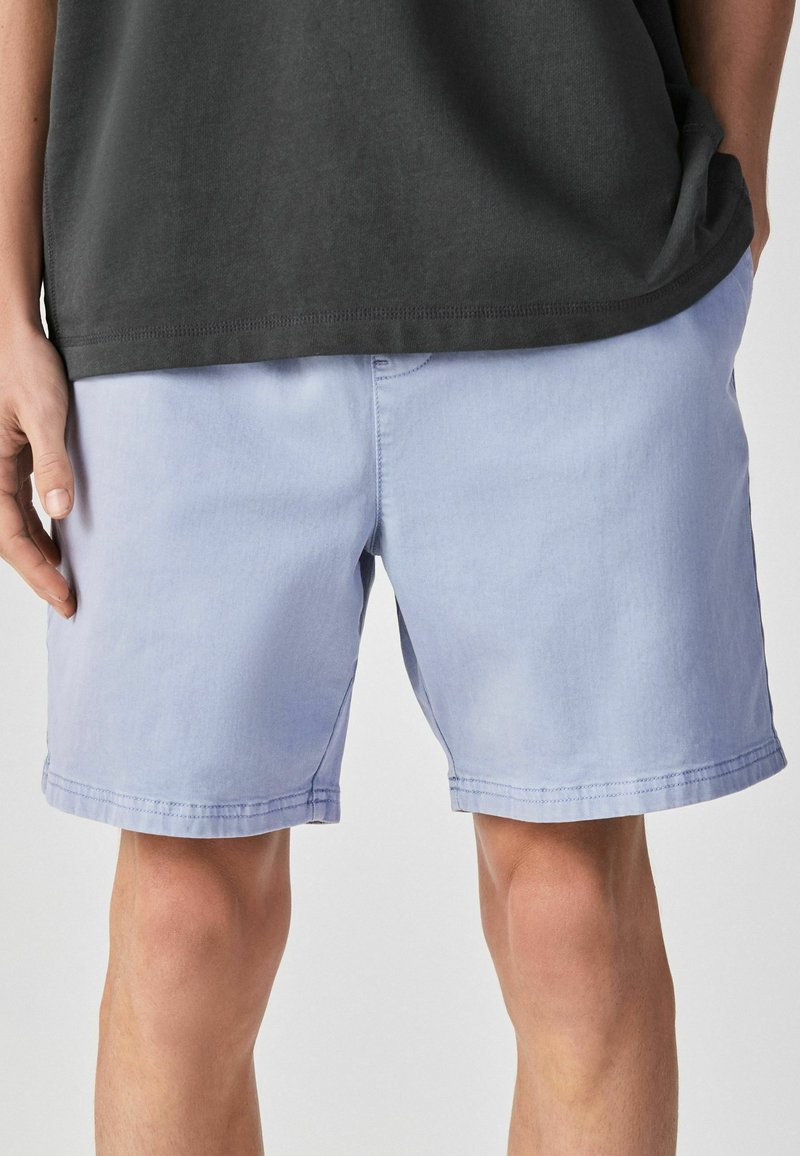 Homme portant un short bleu clair jusqu'aux genoux et une chemise noire, une main dans la poche, sur un fond uni.