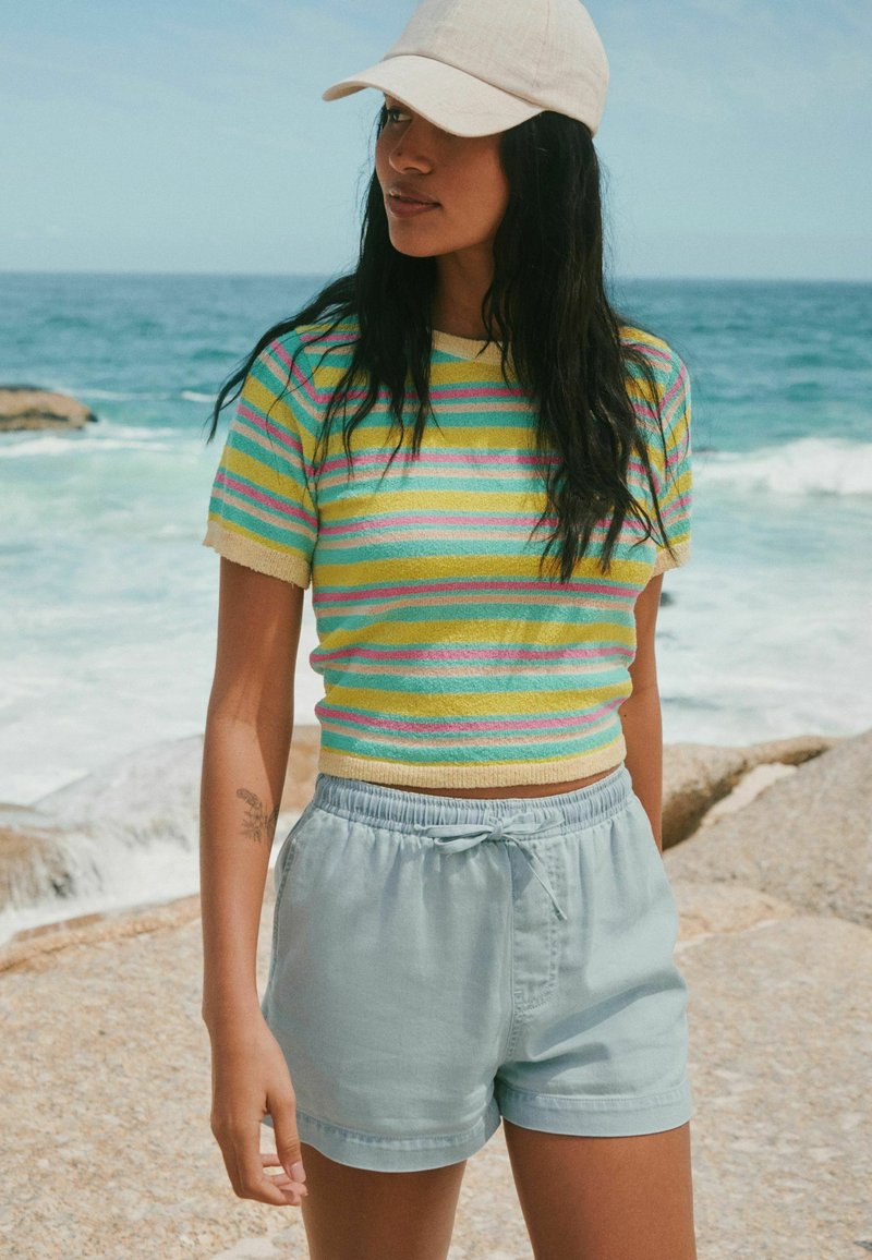 Femme portant une casquette beige, un haut à manches courtes rayé et un short bleu clair, debout sur un rivage rocheux avec des vagues de l'océan en arrière-plan.