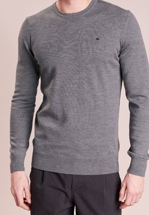 Hombre vistiendo un suéter gris ajustado de manga larga con un pequeño logo negro en el pecho y pantalones oscuros sobre un fondo liso.
