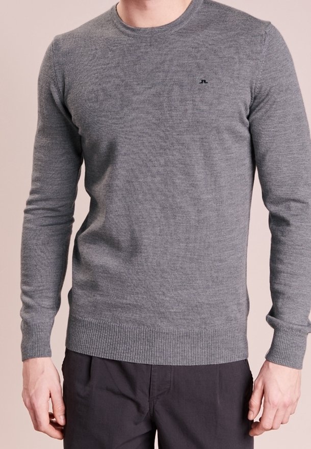 Homme portant un pull gris cintré à manches longues avec un petit logo noir sur la poitrine et un pantalon foncé sur un fond uni.