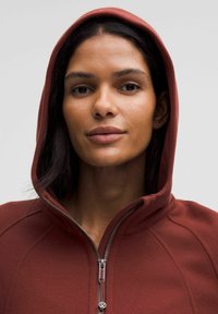 Felpa con zip di colore bordeaux, dotata di cappuccio, tessuto testurizzato e una zip visibile con un accent hardware circolare. Primo piano del collo e del viso.