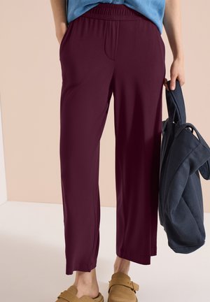 Personne portant un pantalon ample grenat et des sandales beige à enfiler, tenant un sac en tissu bleu foncé dans sa main droite.