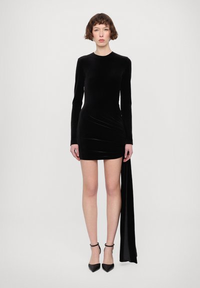 Schwarzes Samtkleid mit langen Ärmeln und asymmetrischem Saum, mit einem drapierten Seitendetail und figurbetonter Silhouette. Schwarze High Heels.