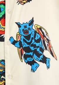 Chemise à manches courtes en tissu beige clair avec un imprimé graphique d'une créature ailée bleue. Les détails comprennent des caractéristiques dessinées et une fermeture à boutons.