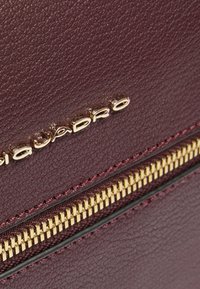 Bolso de cuero burdeos con superficie texturizada, con cremallera de tono dorado y logo de la marca grabado en letras mayúsculas.