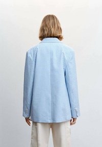 Blazer a scacchi blu e bianchi, design strutturato con colletto a punte, maniche lunghe e polsini con bottoni, abbinato a pantaloni di colore chiaro.