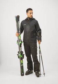 Giacca da sci nera impermeabile con cerniere, pantaloni neri e scarponi da sci neri. Tiene sci con dettagli verdi e bastoni da sci neri.