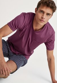 Lila Kurzarm-T-Shirt mit einer Tasche, kombiniert mit gemusterten Shorts in Blau, Weiß und Rot. Der Stoff wirkt glatt und leicht.