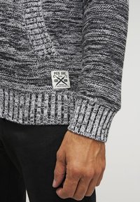 Stickad tröja med svarta och grå horisontella ränder, texturerade ribbade manschetter och en vit etikett med "PIER ONE" på nederkanten.