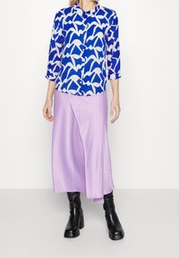 Blusa a maniche tre quarti a fantasia blu e bianca con bottoni, abbinata a una gonna avvolgente in satin lilla e stivaletti neri.