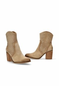 Botines de ante beige con diseño perforado, puntera puntiaguda y tacón apilado. Presentan una superficie texturizada y cortes abiertos en los laterales.