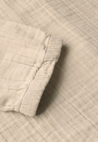 Beiger Musselin-Stoff mit strukturiertem Finish und gerafftem Ärmel. Die weiche, leichte Qualität des Stoffes zeigt sich in den Falten.