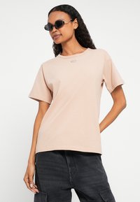 Gaiši rozā kokvilnas T-krekls ar apaļu apkakli, īsām piedurknēm un mazu sudraba krāsas logotipu uz krūtīm, kopā ar melnām kauču biksēm.