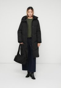 Ung kvinde iført en lang sort dunfrakke, grøn sweater, vide jeans, sorte støvler, mens hun holder en sort håndtaske, stående foran en ensfarvet væg.