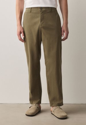 Man wearing olive green straight-leg pants and beige slip-on shoes standing against a plain wall background.