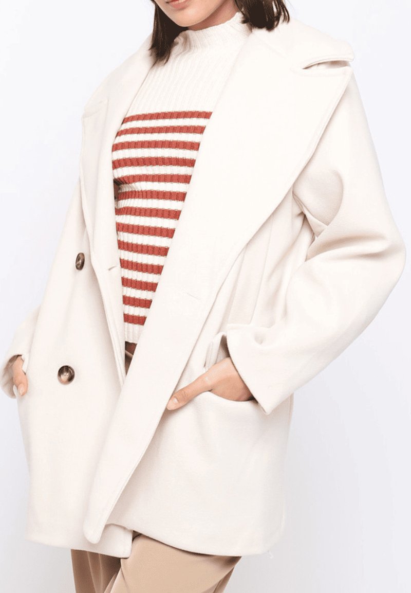 Abrigo de color crema de doble botonadura con grandes solapas, dos bolsillos delanteros y una textura suave, superpuesto sobre un suéter a rayas rojas y blancas.