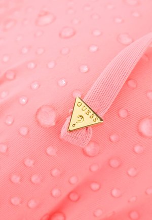 Tela rosa con textura de puntos en relieve y gotas de agua, con un logo triangular dorado de Guess adherido a una banda elástica rosa.