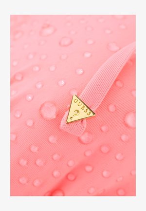 Tela rosa con textura de puntos en relieve y gotas de agua, con un logo triangular dorado de Guess adherido a una banda elástica rosa.