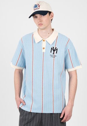 Mand iført en lyseblå strikket polo med røde lodrette striber og cremefarvet krave, hvid kasket og mørke bukser med nålestriber, der står med hænderne i lommerne.