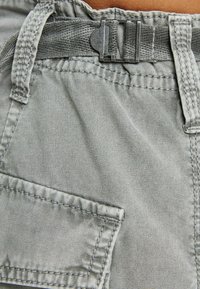 Pantaloni corti in tessuto grigio con una finitura strutturata, dotati di una fibbia in metallo e cuciture visibili. Include un dettaglio con tasca a toppa.