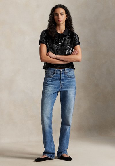 Črna majica s kratkimi rokavi in ovratnikom, okrašena z bleščicami, kombinirana z svetlo modrimi kavbojkami s širokimi hlačnicami in črnimi balerinkami. Preprost, teksturiran dizajn.