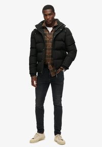 Czarna kurtka puchowa z kapturem, koszula w kratę pod spodem, ciemne dżinsy i beżowe trampki. Posiada zamek błyskawiczny i elastyczne mankiety.