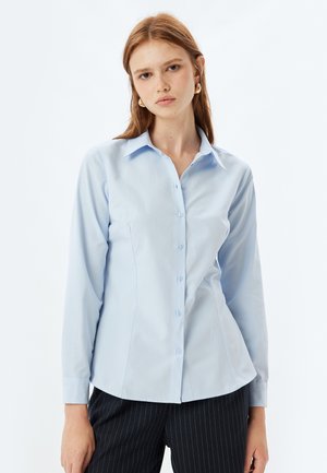 Camisa abotonada azul clara con mangas largas, cuello puntiagudo y un ajuste entallado. Textura suave con costuras sutiles.