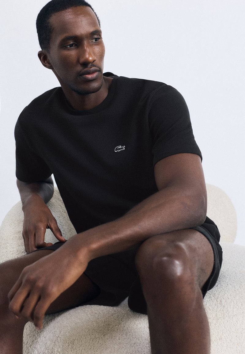 Jovem vestido com uma t-shirt preta da Lacoste e calções, sentado numa cadeira branca texturizada, olhando para a sua esquerda.