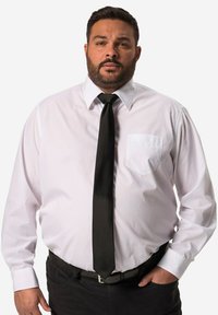 Camicia bianca con maniche lunghe, colletto classico, taschino sul petto; abbinata a una cravatta nera unita e una cintura nera.