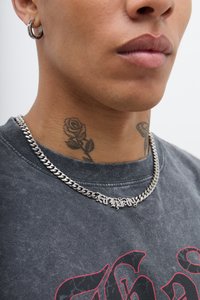 Colar de corrente prateada com uma placa de identificação, usado sobre uma t-shirt preta desbotada com gráficos vermelhos, complementado por um brinco de argola prateado.
