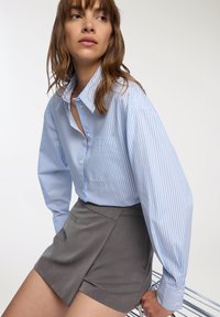 Camicia a righe blu e bianca con bottoni, maniche lunghe e taschino sul petto, abbinata a pantaloni corti grigi tailored. Design semplice e strutturato.