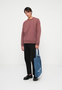 Sudadera color burdeos con cuello redondo y mangas largas, combinada con pantalones negros. Lleva una bolsa de mezclilla azul con texto blanco. Zapatos negros.