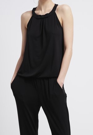 Frau trägt schwarzen ärmellosen Jumpsuit mit Neckholder, Hände in den Taschen, vor einfachem Hintergrund.