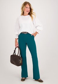 Vrouw met lang blond haar, gehuld in een witte blouse, teal pantalons, en luipaardprint flats, met een donkerbruine handtas in de hand, staande met haar hand in de zak.