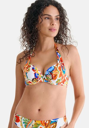 Femme aux cheveux bouclés portant un bikini à bretelles fleuri multicolore, regardant légèrement sur le côté contre un fond uni.