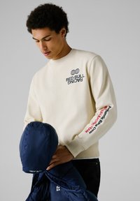 Beige sweatshirt met het "RED BULL RACING" logo op de borst en mouw, gecombineerd met een blauwe pufferjack. Soepele textuur, casual pasvorm.
