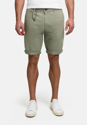 Man draagt opgerolde olijfgroene korte broek, witte sneakers en een wit overhemd, staat met ontspannen armen langs zijn zij.
