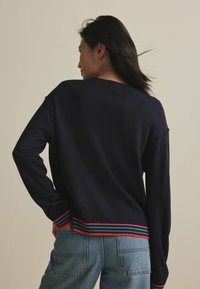Marinblå tröja med långt ärm, ribbat halsringning och muddar, med en flerfärgad rand i grön, rosa och orange i nederkanten. Bärs tillsammans med ljusblå jeans.