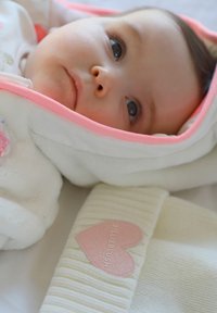 Baby gewikkeld in een witte deken met een roze rand, liggend naast een wit gebreid item met een roze hartlabel met de tekst "Billieblush."
