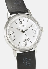 Timex-Uhr mit silbernem Zifferblatt, schwarzem Lederarmband, zeigt Snoopy-Illustrationen, Edelstahlgehäuse und traditionelle Stundenmarkierungen.