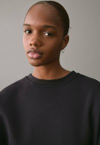 Svart sweatshirt med rund halsringning, ribbade muddändar och en mjuk textur. Modellen har kort hår och små hoops. Enkla bakgrund.