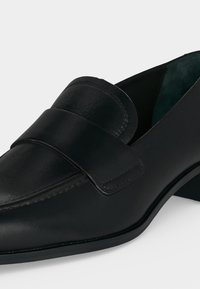 Mocassin femme en cuir noir avec un petit talon bloc et un bout arrondi, avec une large bride sur le dessus.