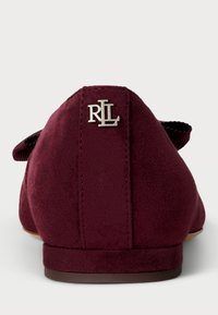 Burgundy mockaläderskor med en dekorativ rosett, platt sula och en metallisk "RL"-logotyp vid hälen. Ytan är slät med en matt finish.