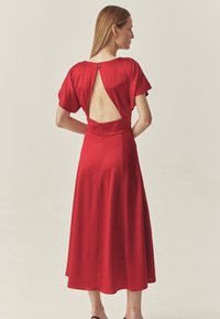 Robe midi en satin rouge avec manches courtes évasées, dos découpé et fermeture par boutons, présentant une texture lisse et une silhouette fluide.