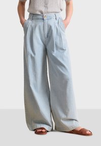 Jeans de pierna ancha de color azul claro hechos de mezclilla, con una cintura alta, pliegues y bolsillos laterales. Combinados con sandalias de tiras marrones.