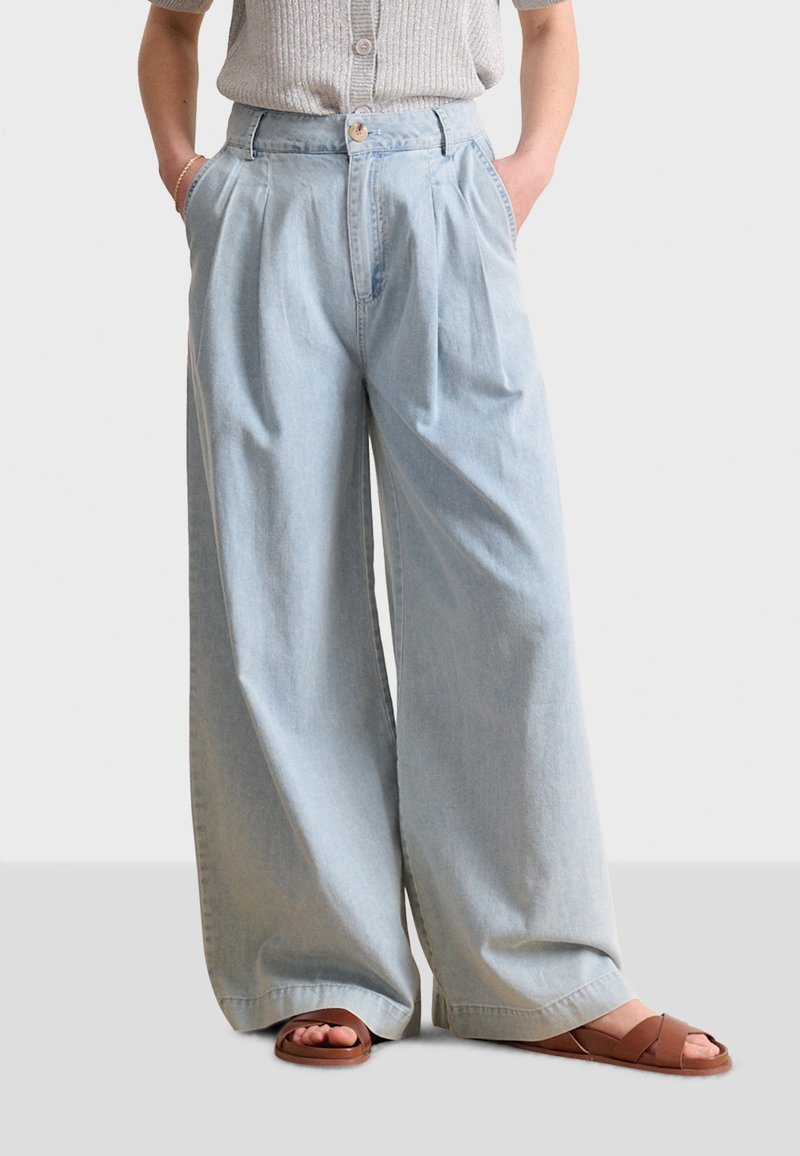Jeans de pierna ancha de color azul claro hechos de mezclilla, con una cintura alta, pliegues y bolsillos laterales. Combinados con sandalias de tiras marrones.