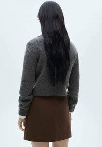 Maglione grigio corto con maniche lunghe, texture lavorata a maglia, abbinato a una gonna marrone. La vista mostra il retro con lunghi capelli scuri.