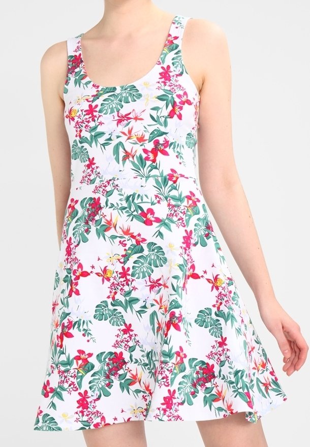 Femme portant une robe blanche sans manches avec des feuilles vertes et un imprimé floral rose, debout devant un fond clair uni.