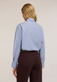 Jasnoniebieska koszula w paski z brązowymi akcentami, z długimi rękawami i kołnierzykiem, noszona z ciemnobrązowymi spodniami. Posiada mankiet zapinany na guzik.