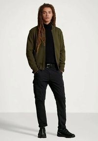 Jaunas vyras su ilgamis dredais stovi su rankomis kišenėse, vilkėdamas alyvuogių žalios spalvos bomberio tipo striukę, juodą megztinį su aukštu kaklu, juodas krovinių kelnes ir juodas sportines batus.